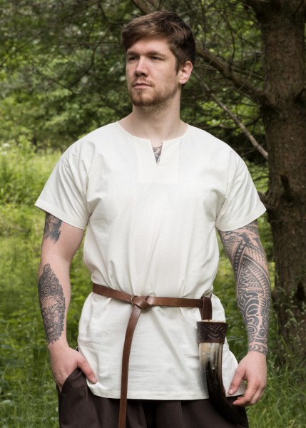 Die einfache Mittelalter-Tunika Sigmund in Naturtönen, kurzarm, wird von einem Mann mit Bart und Tätowierungen im Freien getragen. Die Tunika hat einen geraden Schnitt mit einem Rundhalsausschnitt und besteht aus leichtem Stoff.